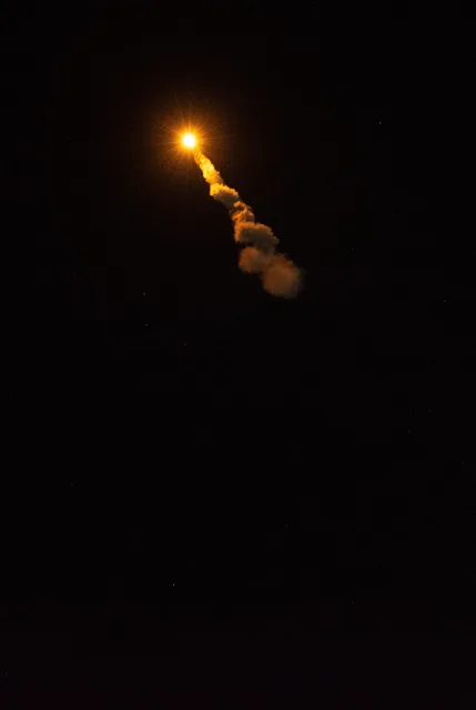A small flame and trail of smoke from a rocket rising into a dark sky.